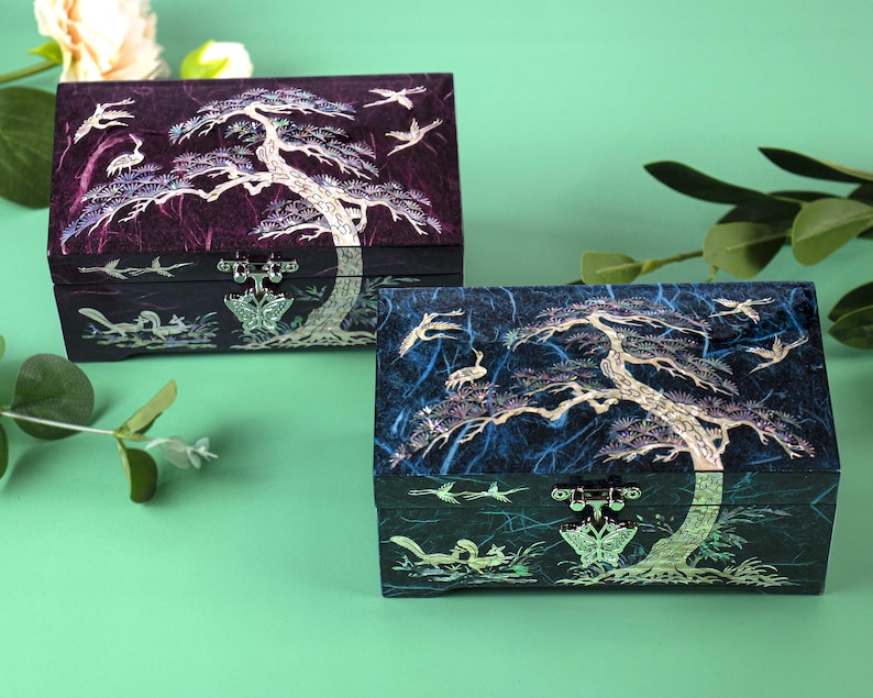 K&ouml;nnte beinhalten: Zwei dekorative Boxen mit aufwendigen Baum- und Vogelmotiven. Die Boxen sind rechteckig mit einem dunkelvioletten und blauen Hintergrund. Der Baum und die V&ouml;gel sind in einer helleren Farbe gehalten, und die Boxen haben einen Schmetterlingsverschluss.