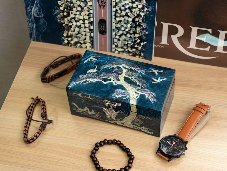 K&ouml;nnte beinhalten: Eine Sammlung von Accessoires, darunter eine tealfarbene Schmuckschatulle mit einem Baum- und Vogeldesign, eine braune Lederuhr und mehrere Armb&auml;nder. Die Uhr hat ein schwarzes Zifferblatt und ein braunes Lederarmband.