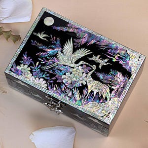 以下が含まれることがあります： 黒い長方形のジュエリーボックス。真珠母貝の象嵌デザイン。鶴のモチーフと花や鳥のディテールが特徴です。シルバー色の留め金と、上部に月のデザインがあります。