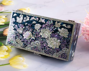 May include: A rectangular clutch bag featuring a dark teal lacquered surface inlaid with iridescent mother-of-pearl floral patterns and butterflies. The edges are framed with a silver-toned metallic border and a geometric Greek key pattern.
