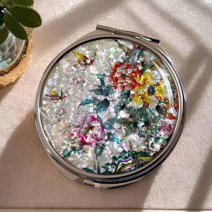 May include: A silver compact mirror with a floral design. The mirror has a white background with pink, yellow, and blue flowers and butterflies.
