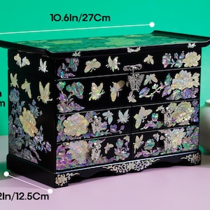 Pode incluir: Uma caixa de joias preta com incrusta&ccedil;&otilde;es de madrep&eacute;rola iridescente de borboletas e flores, com uma tampa superior e quatro gavetas com puxadores de metal em forma de borboleta. Mede 27 cm de largura, 17 cm de altura e 12,5 cm de profundidade.