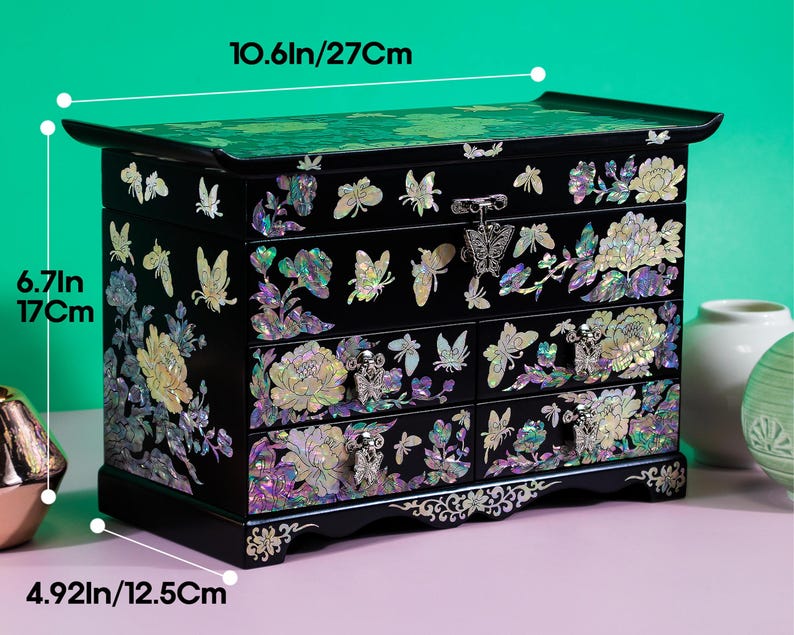 Pode incluir: Uma caixa de joias preta com incrusta&ccedil;&otilde;es de madrep&eacute;rola iridescente de borboletas e flores, com uma tampa superior e quatro gavetas com puxadores de metal em forma de borboleta. Mede 27 cm de largura, 17 cm de altura e 12,5 cm de profundidade.