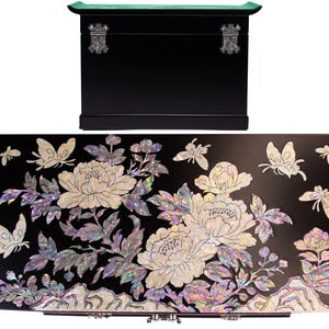 Pode incluir: Uma caixa de armazenamento retangular preta com tampa curva e fechos de metal prateado. O painel frontal apresenta uma incrusta&ccedil;&atilde;o complexa de madrep&eacute;rola iridescente representando flores de pe&ocirc;nia, ramos folhosos e v&aacute;rias borboletas.