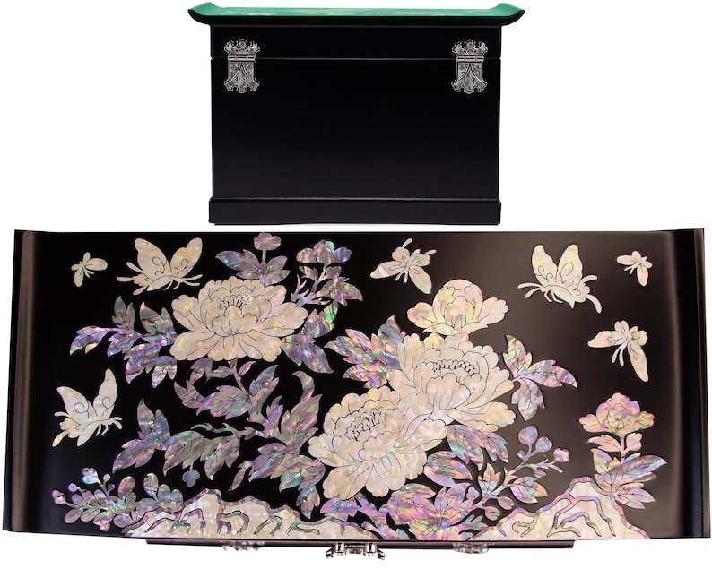 Pode incluir: Uma caixa de armazenamento retangular preta com tampa curva e fechos de metal prateado. O painel frontal apresenta uma incrusta&ccedil;&atilde;o complexa de madrep&eacute;rola iridescente representando flores de pe&ocirc;nia, ramos folhosos e v&aacute;rias borboletas.