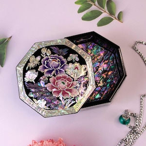 May include: A black, octagonal jewellery box with a floral mother-of-pearl inlay, open to reveal a shimmering interior. The lid displays a pink and purple peony design. A silver necklace with a green pendant is visible.