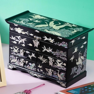 May include: Black jewelry box with a green top and multiple drawers. The box is decorated with mother-of-pearl inlay of cranes and other birds. The box has a butterfly-shaped clasp and is on a pink surface.
