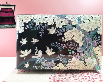 May include: Black jewelry box with a glossy finish, adorned with mother-of-pearl inlay. The design features white flowers, branches, and birds in flight. The box has a hinged lid and a velvet-lined interior.