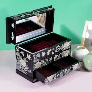 Pode incluir: Uma caixa de joias preta com tampa articulada, espelho interior e compartimento forrado a veludo. O exterior &eacute; decorado com incrusta&ccedil;&otilde;es de madrep&eacute;rola iridescente de borboletas e flores, incluindo duas gavetas extra&iacute;veis.