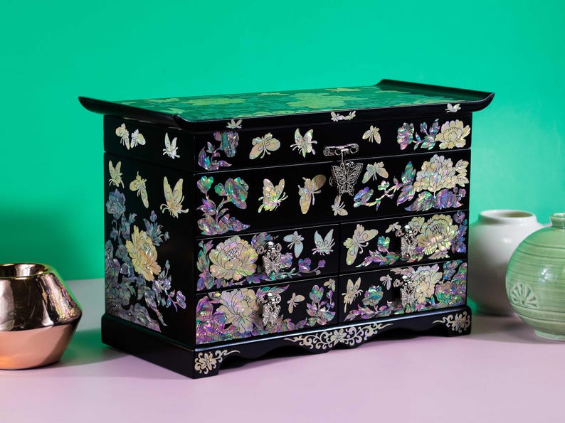 Pode incluir: Um porta-joias de madeira preta com incrusta&ccedil;&otilde;es de madrep&eacute;rola representando borboletas e motivos florais. O ba&uacute; inclui uma tampa superior e quatro gavetas, cada uma adornada com puxadores de metal em forma de borboleta e detalhes iridescentes.