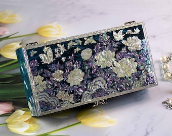 May include: A rectangular clutch bag featuring a dark teal surface inlaid with iridescent mother-of-pearl designs of blooming flowers, butterflies, and stylized clouds. The edges are framed in silver-toned metal with a geometric border pattern.