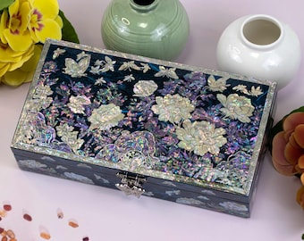 May include: Dark blue rectangular jewellery box with a floral and butterfly design. The box is decorated with iridescent mother-of-pearl inlays, featuring large white flowers and delicate butterflies. It has a silver-coloured clasp.