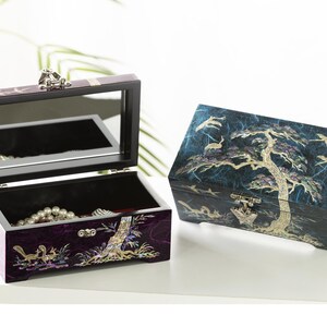 K&ouml;nnte beinhalten: Zwei dekorative Schmuckk&auml;stchen. Eines ist ge&ouml;ffnet und zeigt einen Spiegel und Schmuck im Inneren. Die andere Box ist geschlossen. Beide Boxen weisen aufwendige Designs mit B&auml;umen, V&ouml;geln und anderen Naturelementen in einem dunkelvioletten und blauen Farbschema auf.