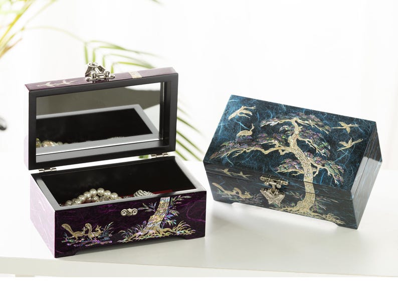 K&ouml;nnte beinhalten: Zwei dekorative Schmuckk&auml;stchen. Eines ist ge&ouml;ffnet und zeigt einen Spiegel und Schmuck im Inneren. Die andere Box ist geschlossen. Beide Boxen weisen aufwendige Designs mit B&auml;umen, V&ouml;geln und anderen Naturelementen in einem dunkelvioletten und blauen Farbschema auf.