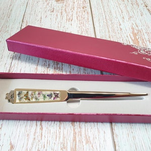 May include: A gold letter opener with a white handle decorated with colorful butterflies. The letter opener is in a red velvet box with a white floral design.