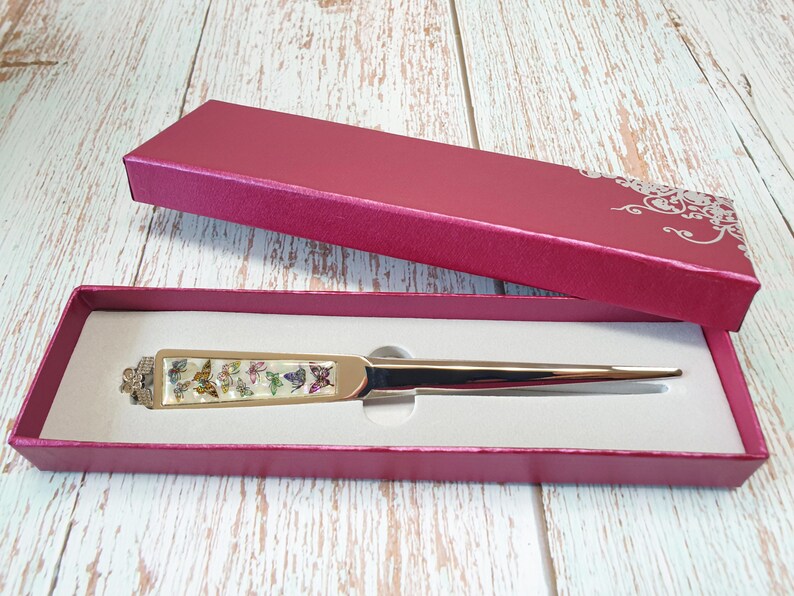 May include: A gold letter opener with a white handle decorated with colorful butterflies. The letter opener is in a red velvet box with a white floral design.