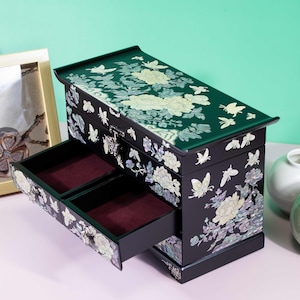 Pode incluir: Uma caixa de joias retangular preta com designs intrincados de incrusta&ccedil;&otilde;es de madrep&eacute;rola de flores desabrochando e borboletas. A caixa inclui uma tampa articulada e duas gavetas com forro de veludo vermelho escuro e fecho.