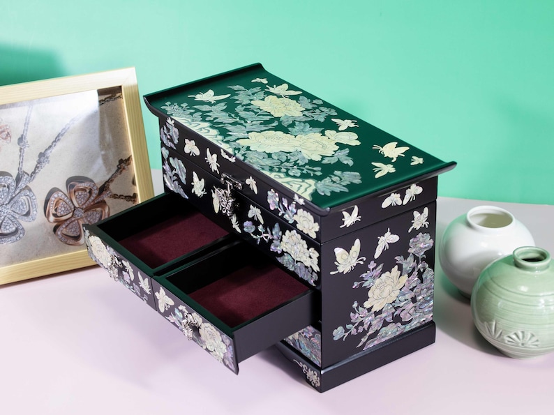 Pode incluir: Uma caixa de joias retangular preta com designs intrincados de incrusta&ccedil;&otilde;es de madrep&eacute;rola de flores desabrochando e borboletas. A caixa inclui uma tampa articulada e duas gavetas com forro de veludo vermelho escuro e fecho.