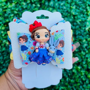 May include: A blue and white bow with a small doll of Snow White holding a bunny. The doll has brown hair, a red bow, and a blue dress. The bow is decorated with flowers and leaves.