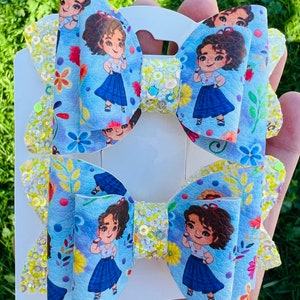 May include: Two blue and white bow hair clips with a floral pattern and a cartoon character of a girl with brown hair and a blue dress. The bows are decorated with yellow glitter.