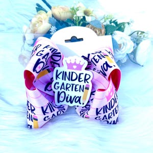 May include: Pink hair bow with a white and black "Kindergarten Diva" design with a crown and pencil. The bow is made of ribbon with a repeating pattern of "Kindergarten Diva" and pencils.