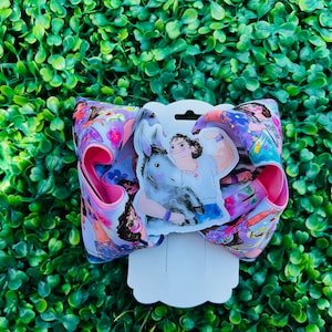 May include: A pink hair bow with a colorful print featuring a cartoon woman and a donkey. The print includes flowers and stars. The bow is attached to a white card with a hole in the center.