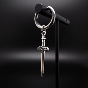 May include: A silver hoop earring with a sword charm. The sword has intricate details and a pointed tip.