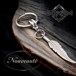 Può includere: Un orecchino color argento con un ciondolo a forma di piuma. L'orecchino ha un design a cerchio con una piccola catena che si collega alla piuma. La parola "Nouveauté" è in un ovale dorato. Il marchio "GORRM" è in bianco.