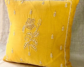 May include: A square mustard yellow throw pillow cover featuring white embroidered floral motifs and a central hibiscus flower design. The edges are defined by a delicate white stitched border, with small leaf patterns scattered across the surface.