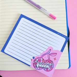 May include: A blue and white lined notepad with a pink cherry sticker that says "dancing queen" on it. The sticker has a disco ball design and glitter accents.