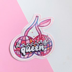 May include: A pink and red glitter sticker with two cherries and the text "dancing queen" in a black, cursive font.