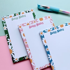 May include: Three rectangular notepads with daisy borders in green, pink, and blue. Each notepad has the text "Daisy Notes" in a coordinating color. A multi-colored pen rests on a light blue surface.