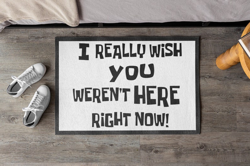 Puede incluir: Felpudo blanco con borde negro y el texto "I REALLY WISH YOU WEREN'T HERE RIGHT NOW!" en letras negras en negrita. Dos pares de zapatillas blancas est&aacute;n en el suelo de madera junto a la alfombra.