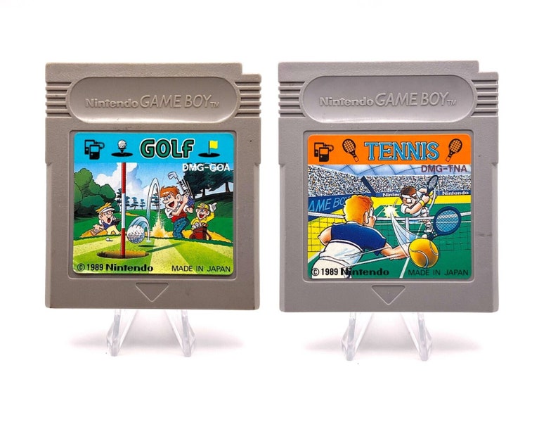K&ouml;nnte beinhalten: Zwei Vintage-Game-Boy-Cartridges, eine f&uuml;r Golf und eine f&uuml;r Tennis. Beide Cartridges sind grau mit bunten Etiketten, die Illustrationen der Spiele zeigen. Der Text auf den Etiketten lautet "Nintendo GAME BOY", "GOLF", "DMG-GOA", "TENNIS", "DMG-TNA", "1989 Nintendo" und "MADE IN JAPAN".