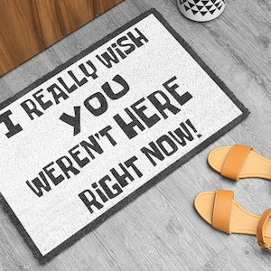 Puede incluir: Felpudo rectangular blanco con texto negro: "I REALLY WISH YOU WEREN'T HERE RIGHT NOW!". Borde gris. Junto a &eacute;l, sandalias color canela con correas en los tobillos. Decoraci&oacute;n para el hogar.