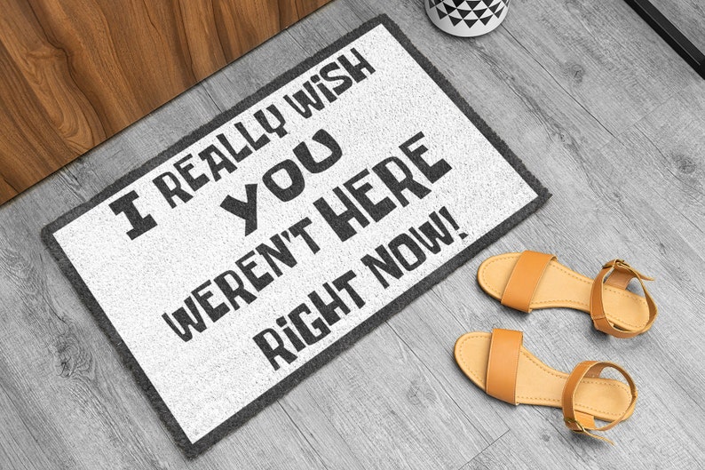 Puede incluir: Felpudo rectangular blanco con texto negro: "I REALLY WISH YOU WEREN'T HERE RIGHT NOW!". Borde gris. Junto a &eacute;l, sandalias color canela con correas en los tobillos. Decoraci&oacute;n para el hogar.