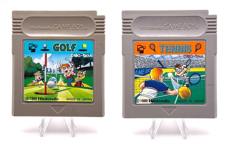 K&ouml;nnte beinhalten: Zwei Vintage Game Boy-Cartridges, eine f&uuml;r Golf und eine f&uuml;r Tennis. Beide Cartridges sind grau mit bunten Etiketten und dem Text "Nintendo GAME BOY" oben. Die Golf-Cartridge zeigt einen gr&uuml;nen Golfplatz mit einem Golfer und einem Ball in der Luft. Die Tennis-Cartridge zeigt einen Tennisplatz mit zwei Spielern und einem Ball in der Luft.