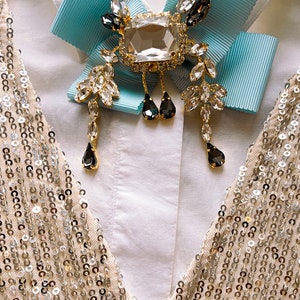 May include: A white shirt with a light blue bow tie and a large crystal brooch with black accents. The brooch has a square-shaped center stone and dangling crystal drops.
