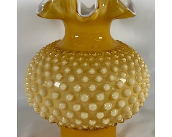 Pode incluir: Um vaso de vidro redondo com uma cor âmbar vibrante e um padrão de textura hobnail com pontos brancos em relevo. O vaso tem uma abertura larga, ondulada e franzida no topo, terminando numa base âmbar lisa e sólida na parte inferior.