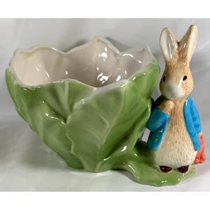 May include: A ceramic planter shaped like a green cabbage leaf bowl with a standing brown rabbit figure attached to the side. The rabbit has white fur on its face and chest, wears a blue jacket, and holds an orange carrot in its right hand.