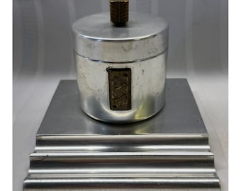 Puede incluir: Un recipiente cilíndrico de metal plateado con tapa, que presenta un pequeño pomo rectangular de color latón en la parte superior y una placa rectangular a juego en el lateral. El recipiente descansa sobre una base cuadrada de metal plateado.