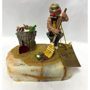 May include: A figurine of a clown with a red nose and hat, holding a yellow broom, standing on a stone base next to a gray trash can. A red dustpan, green bottles, and gold glitter are on the base. A tag reads Creative Concept By Ronald Lee.