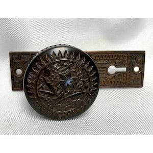 May include: A dark bronze-toned metal door knob and rectangular backplate set. The circular knob features a raised floral motif surrounded by a sunburst-style edge. The rectangular backplate includes a keyhole and decorative etched patterns.
