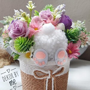 May include: A white bunny rabbit figurine with pink feet sits in a woven basket filled with a floral arrangement of purple, pink, and white flowers. The basket is decorated with a white ribbon and the text "DEKOR BY ME" is visible on the side.