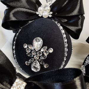 Puede incluir: Adorno navide&ntilde;o de terciopelo negro con un dise&ntilde;o de diamantes de imitaci&oacute;n plateados y un lazo de sat&eacute;n negro con detalles de perlas. El adorno tiene el texto "DEKOR BY ME" en una etiqueta blanca.