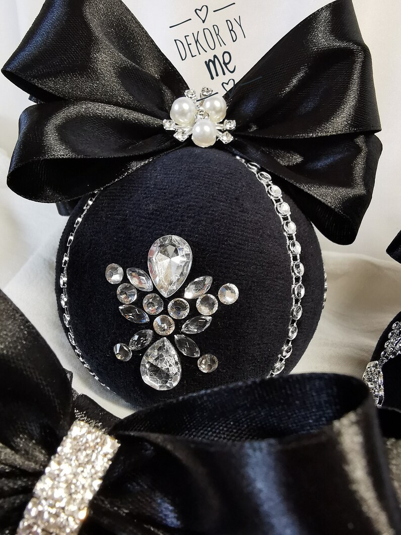Puede incluir: Adorno navide&ntilde;o de terciopelo negro con un dise&ntilde;o de diamantes de imitaci&oacute;n plateados y un lazo de sat&eacute;n negro con detalles de perlas. El adorno tiene el texto "DEKOR BY ME" en una etiqueta blanca.