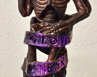 May include: A bronze-colored skeleton couple statue embracing, with a purple ribbon wrapped around them that reads "LOVE NEVER DIES." One skeleton has a purple flower on its head. The statue is on a dark base.