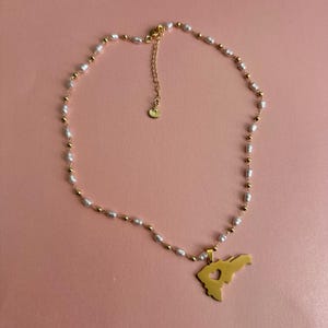May include: A gold-colored necklace with a pendant shaped like a map of a region, featuring a heart cutout. The necklace has a chain of small white pearls and gold-colored beads. The background is a solid pink color.
