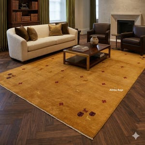 Op de afbeelding: Een groot rechthoekig vloerkleed in een goudgele tint met kleine, verspreide geborduurde motieven van dieren en bloemvormen in donkerrood en goud, met een witte franjerand en de tekst Alihba Rugs op het oppervlak gedrukt.