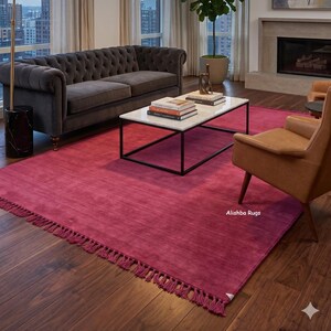 May include: A large, rectangular, solid magenta area rug featuring a short, knotted fringe along the edges. The rug is positioned on a dark wood floor and displays the text Alishba Rugs in the lower right corner of the fabric surface.